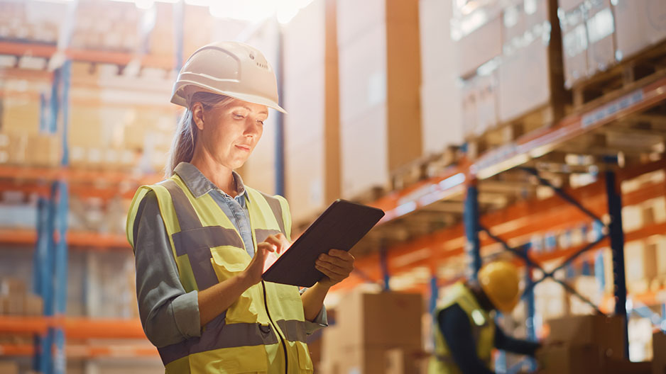 Professional Female Worker Wearing Hard Hat Checks Stock and Inventory with Digital Tablet Computer in the Retail Warehouse